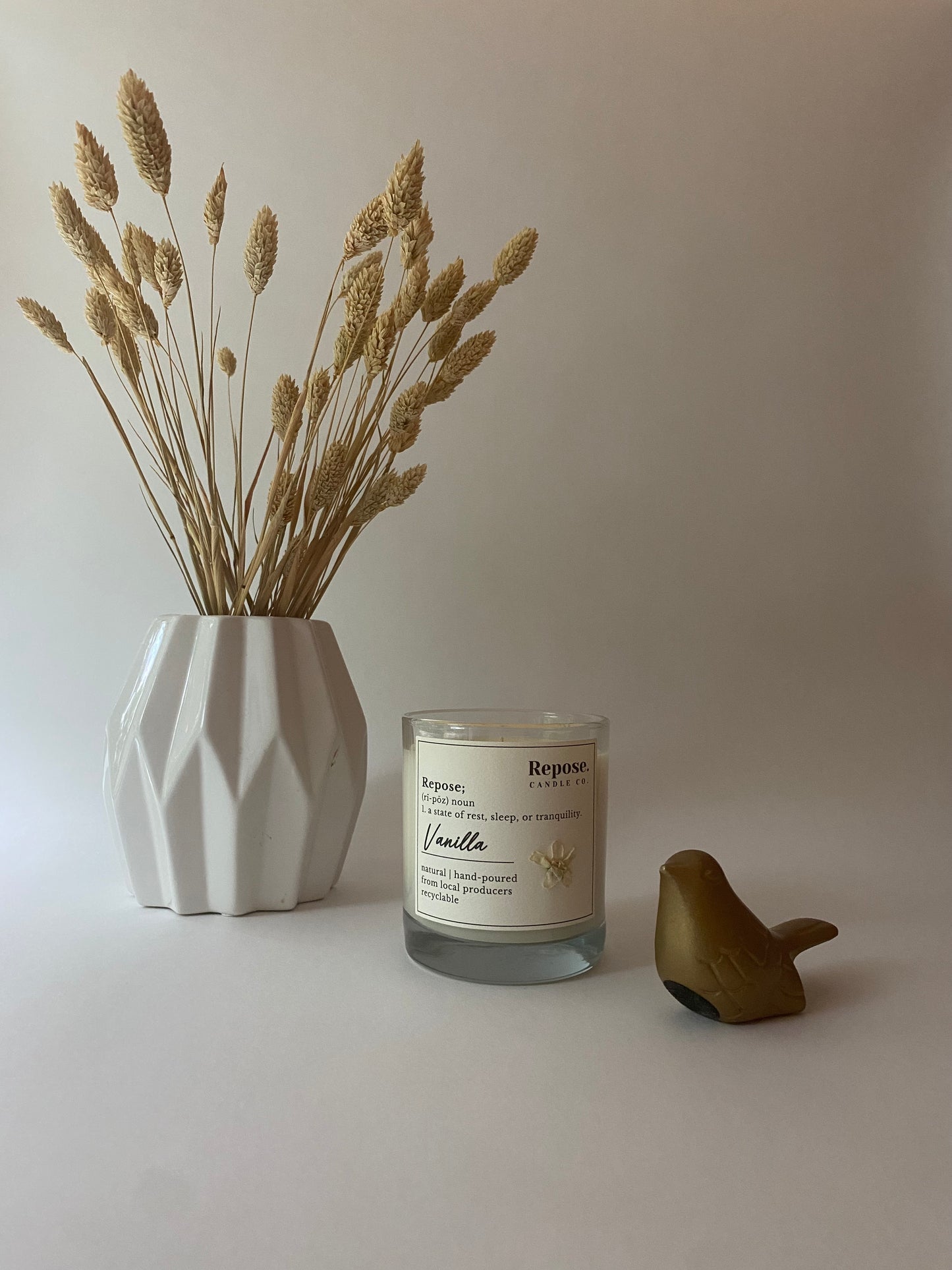 A vanilla candle, a bird figurine, and a white vase with wheat.