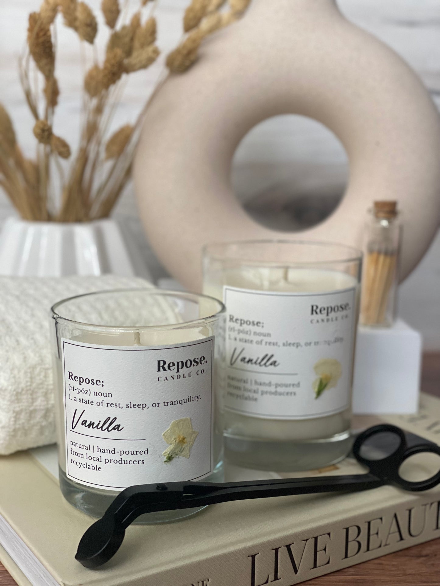 Two Vanilla Candles with wick trimmer, matchesm white towel, vase of wheat and circular cream vase.