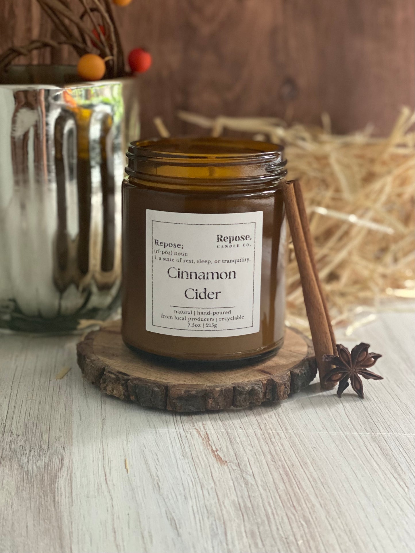 A cinnamon cider candle next to a cinnamon stick and a clove, in front of hay and a silver vase.