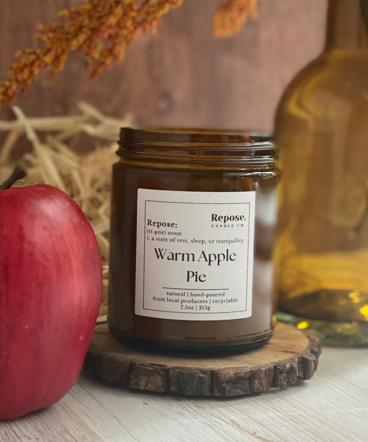 Warm apple pie candle next to an apple and in front of hay and a vase.